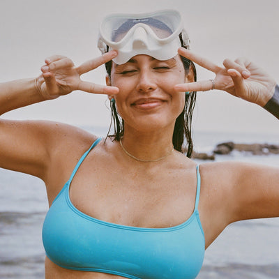 Woman in a blue bikini top with goggles on her head, making peace signs with her fingers, against a beach background.